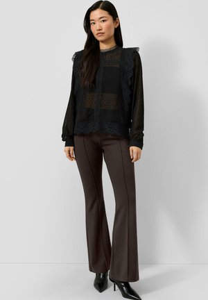 Femme debout portant un chemisier en dentelle noire transparente avec des volants et un pantalon évasé marron foncé avec des bottes noires à talons pointus.