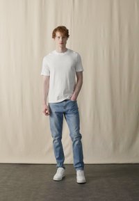 Hvit t-skjorte med korte ermer sammen med lyseblå jeans. Modellen har på seg hvite joggesko. Glatt stoff og avslappet passform. Nøytral bakgrunn.