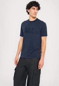 Tamsiai mėlynas trumpų rankovių marškinėlis su apykakle, priekyje juodai išspausdinta toninė "EA7 EMPORIO ARMANI" logotipo gamyba.