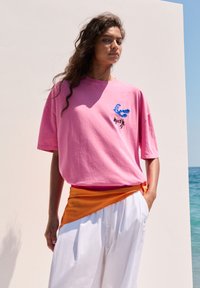 Pink oversized t-shirt med et blåt og sort grafisk design og korte ærmer, parret med hvide vide bukser og et orange taljebånd.