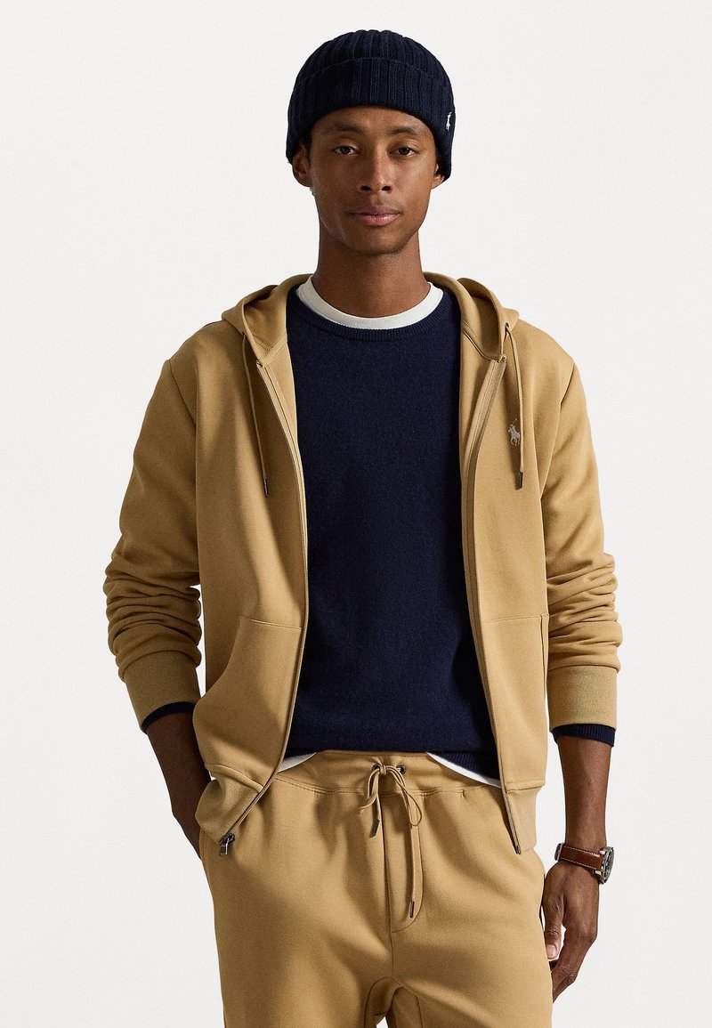 Fleece de capuz bege com fecho na frente e bolsos, por cima de um suéter azul de gola arredondada. Touca azul com nervuras usada e calças bege a condizer com cordão na cintura.