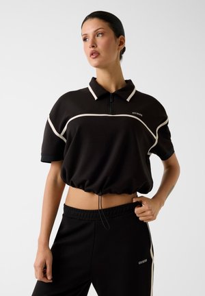 Frau trägt ein schwarzes, kurz geschnittenes Poloshirt mit weißer Paspelierung und passende schwarze Hose, posiert mit einer Hand auf der Hüfte.