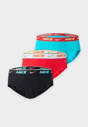Großer, schlanker dunkelhäutiger Mann steht barfuß, trägt schwarze Nike-Slips und schaut seitlich mit einem Arm hinter dem Rücken vor weißem Hintergrund.