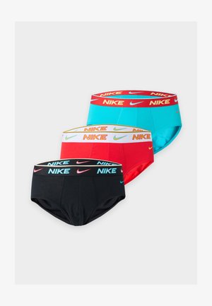 Høj, slank mørkhudet mand står barfodet iført sorte Nike briefs, kigger til siden med en arm bøjet bag ryggen mod en hvid baggrund.
