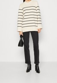 Suéter crema con rayas negras, mangas largas, combinado con jeans negros de pierna recta y botines de tacón. Bolso negro en la mano.