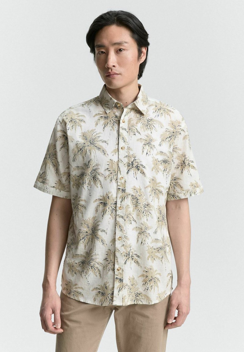Chemise à manches courtes en tissu beige clair avec un motif de palmier en brun foncé. Fermeture à boutons avec un col classique et coupe décontractée.
