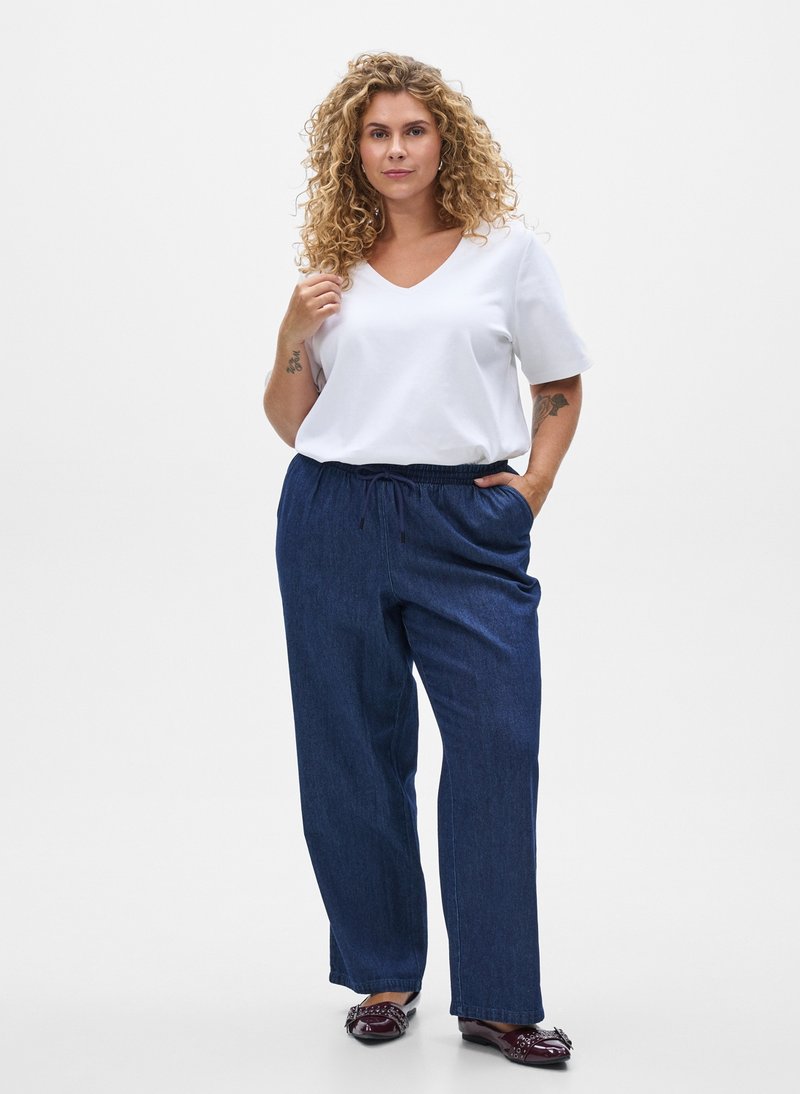 Donna con capelli biondi ricci, che indossa una camicetta bianca a maniche corte, pantaloni larghi blu scuro e scarpe viola scuro, in piedi con una mano in tasca.