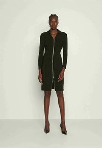 Mulher em pé vestindo um vestido ajustado preto de mangas longas com fecho na frente e sapatos de salto alto pretos de bico fino, contra um fundo claro e simples.