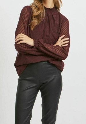 Femme portant un chemisier bordeaux texturé à manches longues avec un motif chevron et un col montant, associée à un pantalon en cuir noir, bras croisés.