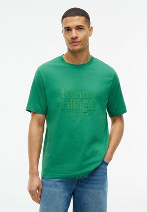 Uomo che indossa una t-shirt verde Tommy Hilfiger e jeans blu, in piedi con una mano in tasca su sfondo bianco.