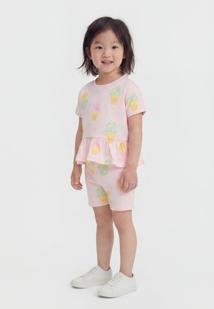 Joven niña de pie, vestida con camiseta rosa de manga corta y pantalones cortos a juego con un patrón colorido de cucuruchos de helado y zapatillas blancas.