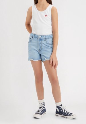 Kind trägt weißes ärmelloses Oberteil mit rotem Logo, hellblaue Jeansshorts, weiße Knöchelsocken und marineblaue High-Top-Sneaker, steht auf weißem Hintergrund.
