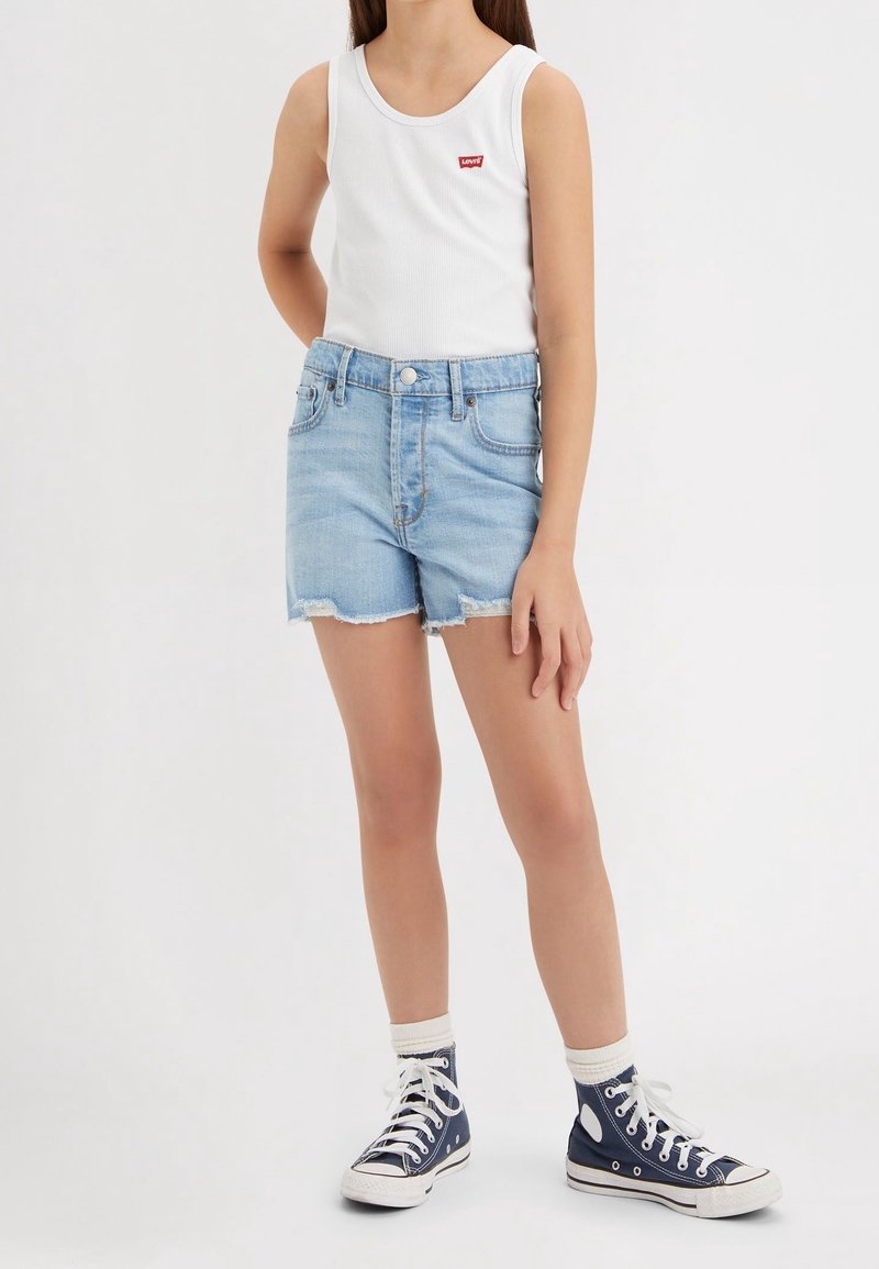 Enfant portant un débardeur blanc avec un logo rouge, un short en denim bleu clair, des chaussettes blanches aux chevilles et des baskets montantes bleu marine, debout sur un fond blanc.
