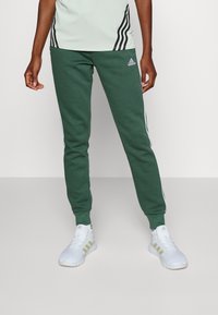 Adidas gröna sweatpants med vita sidostråk, slim fit, tillverkade i mjukt tyg, i kombination med vita sneakers med guldaccenter.