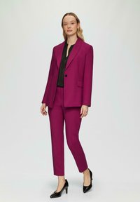 Un costume magenta avec une veste cintrée et des pantalons fuselés, complété par un chemisier noir et des escarpins noirs. Texture de tissu lisse.