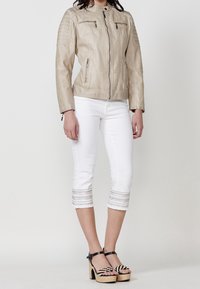 Beżowa skórzana kurtka o dopasowanym kroju, z zamykanymi na zamek kieszeniami i teksturowanymi ramionami, zestawiona z białymi, skróconymi dżinsami z paskowanym wykończeniem.