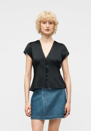 Frau mit kurzen, lockigen blonden Haaren, die eine schwarze V-Ausschnitt-Bluse mit Knöpfen und einen blauen Jeans-Minirock trägt, steht vor einem einfarbigen Hintergrund.