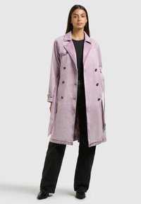 Donna in piedi che indossa un trench doppiopetto viola chiaro sopra una maglietta nera e jeans neri a gamba larga con scarpe nere.