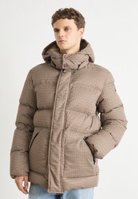 Casaco puffer xadrez em castanho e bege com fecho éclair frontal, gola alta e capuz. Apresenta bolsos laterais e uma textura acolchoada.