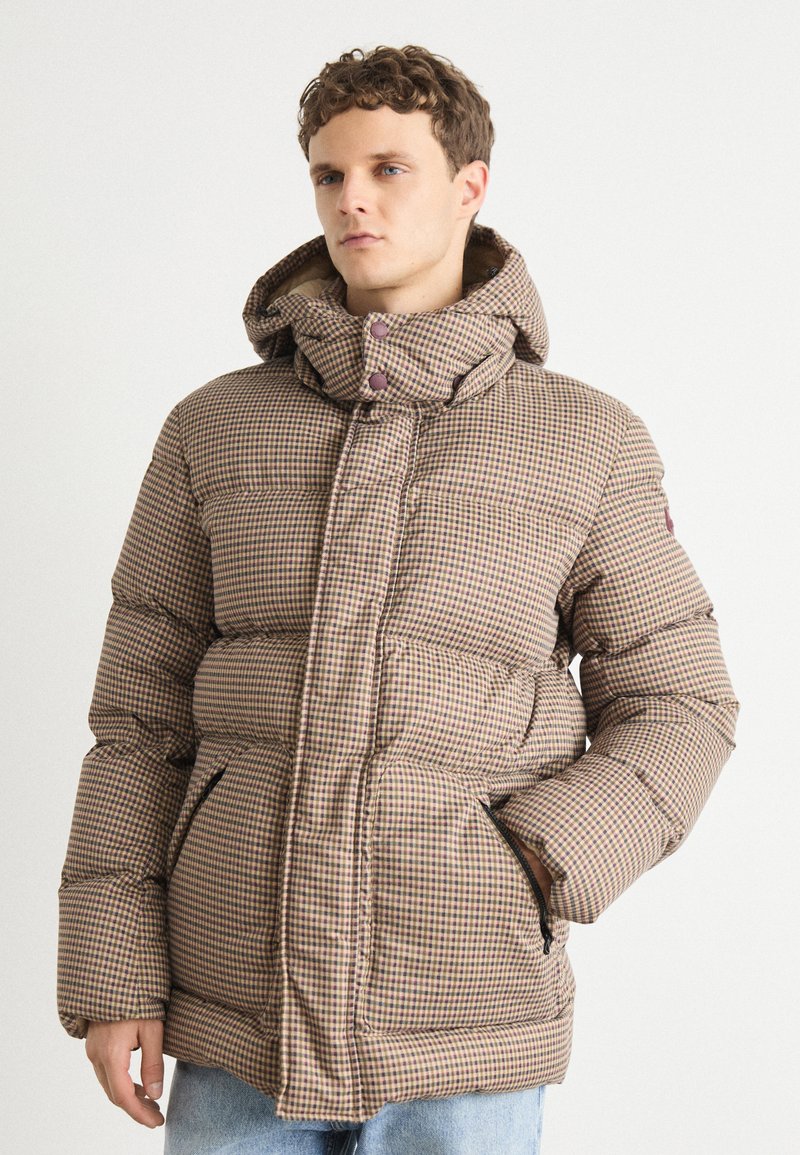 Casaco puffer xadrez em castanho e bege com fecho éclair frontal, gola alta e capuz. Apresenta bolsos laterais e uma textura acolchoada.