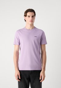 Camiseta de color lavanda claro, hecha de algodón suave, con un cuello redondo y mangas cortas, que presenta un pequeño logo negro en el lado izquierdo del pecho.