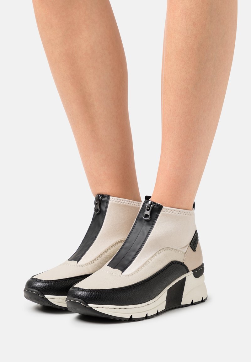 Bottes de cheville beiges et noires avec fermeture éclair, présentant une finition texturée et une semelle compensée, des couleurs contrastantes et un design épuré.