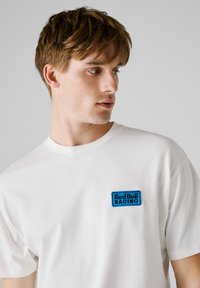 Witte katoenen t-shirt met een blauw geborduurd "Red Bull Racing" logo op de linkerkant van de borst, ronde halslijn en korte mouwen.