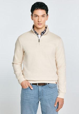 Fiatal férfi bézs színű, negyed cipzáras pulóvert visel kockás ing fölött és világoskék farmert, egyik kezét a zsebében tartva áll.