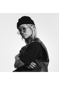 Beanie nero, occhiali rettangolari trasparenti e un gilet nero senza maniche sopra una maglietta. Tatuaggi notevoli su braccia e viso, con uno sfondo in bianco e nero.