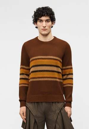 Jeune homme aux cheveux bouclés portant un pull marron en tricot avec des rayures horizontales orange et beige, et un pantalon large à carreaux marron.