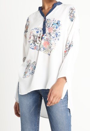 Weiße Langarmbluse mit blauen Blumenmustern und einem dunkelblauen Kragen, kombiniert mit blauen Jeans, getragen von einer stehenden Person.