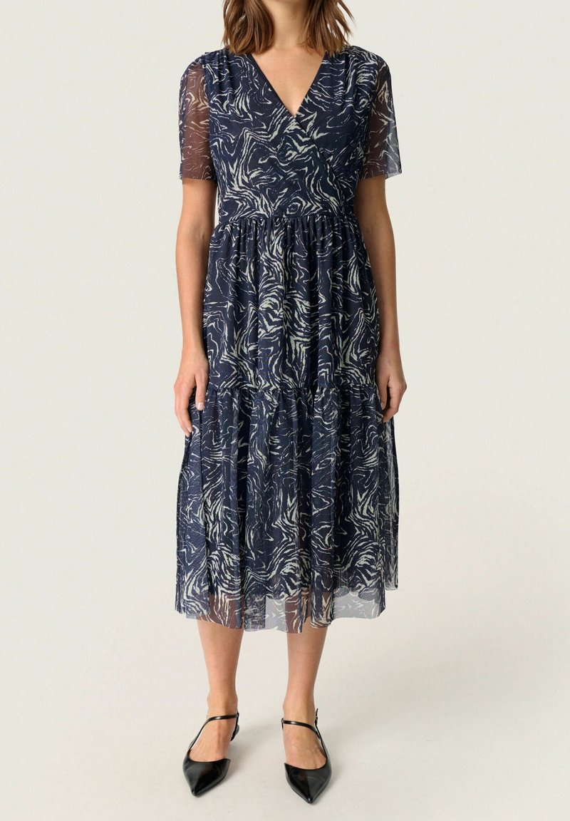 Žena ve středně dlouhých námořnických modrých šatech s bílým abstraktním vzorem, s průhlednými krátkými rukávy, výstřihem do V a černými špičatými T-páskovými botami.