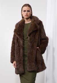 Manteau en fausse fourrure marron avec un large col, au design volumineux et à la texture douce, porté sur une robe verte.