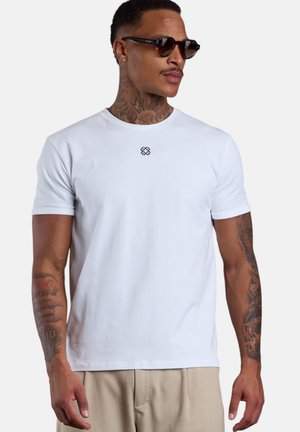 Man with tattoos wearing dark sunglasses, a plain white t-shirt with small black logo at chest, and beige pants, facing slightly right.