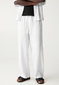 Personne portant un pantalon en lin blanc ample avec une taille à cordon, des sandales noires à boucles, et une chemise blanche partiellement visible sur un haut noir.