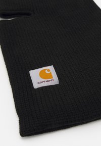 Crna pletena balaclava s teksturiranom površinom, sa pravokutnom bijelom etiketom s narančastim logotipom i tekstom "carhartt".