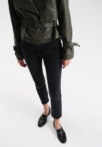 Groene leren jas met brede revers en een ceintuur in de taille, gecombineerd met zwarte cropped jeans en zwarte instappers met gouden hardware details.