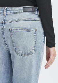Vaqueros de mezclilla azul claro con una etiqueta de marca de cuero negro en la cintura y un bolsillo trasero que presenta un patrón de costura clásico.
