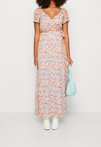 Abito maxi floreale con design avvolgente, maniche a sbuffo corte e vita con laccetti. Sfondo azzurro chiaro con fiori arancioni e rosa. Stivaletti bianchi alla caviglia e borsa azzurro chiaro.