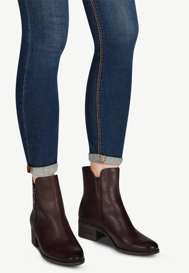 Jambes portant un jean slim bleu foncé retroussé, associé à des bottines en cuir marron foncé avec talons blocs et fermetures éclair latérales.