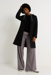 Abrigo negro con capucha y cordones grises, mangas largas y botones. Combinado con pantalones grises de pierna ancha y zapatillas. Fondo liso.