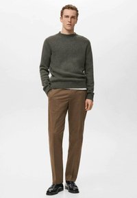 Pull en tricot vert olive avec col rond, associé à un pantalon marron. Le modèle porte des chaussures noires, mettant en valeur une coupe décontractée et une texture apparente.