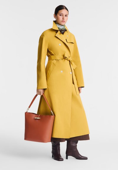 Gelber Trenchcoat mit gebundenem Taillenbund, großen Taschen und silbernen Knöpfen, kombiniert mit einer braunen Handtasche und braunen Stiefeln bis zur Kniehöhe.