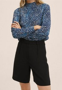 Blouse bleue à manches longues avec un motif blanc éparpillé, associée à un short noir ajusté, présentant un col montant et une coupe droite.