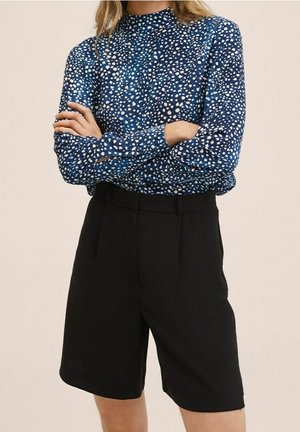 Blusa blu a maniche lunghe con un motivo bianco sparso, abbinata a pantaloni corti neri sartoriali, caratterizzata da un collo alto e una linea dritta.