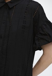 Μαύρο πουκάμισο με κατακόρυφη κεντητή λεπτομέρεια με τρυπούλες και κοντά φουσκωτά μανίκια στο ανώτερο μέρος του κορμού και του βραχίονα ενός ατόμου.