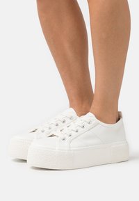 Baskets blanches en toile avec une semelle plateforme en caoutchouc texturé blanc. Comprend des lacets ronds et des détails latéraux subtils.