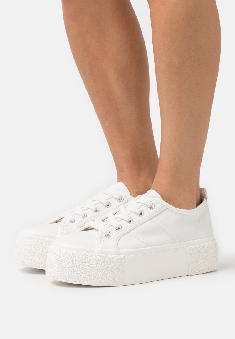 Baskets blanches en toile avec une semelle plateforme en caoutchouc texturé blanc. Comprend des lacets ronds et des détails latéraux subtils.