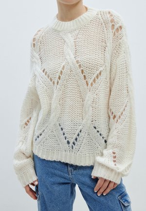Cremefarbener Strickpullover in lockerem Schnitt mit offener Maschenware, versehen mit Rautenmuster und geripptem Ausschnitt sowie Bündchen. Kombiniert mit blauen Jeans.