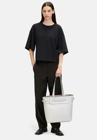 Biała skórzana torba tote o wyrafinowanym kształcie, z podwójnymi uchwytami i subtelnymi detalami. Modelka ma na sobie czarną, oversize'ową koszulkę oraz spodnie w paski.
